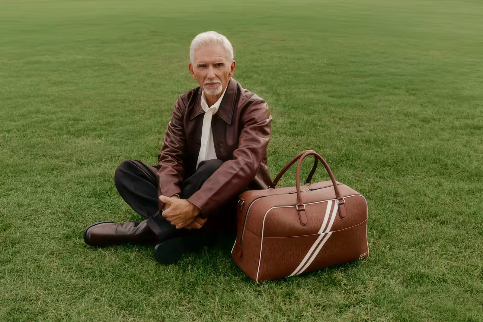 Man in a brown leather jacket sitting on grass beside a Carl Friedrik leather weekender bag